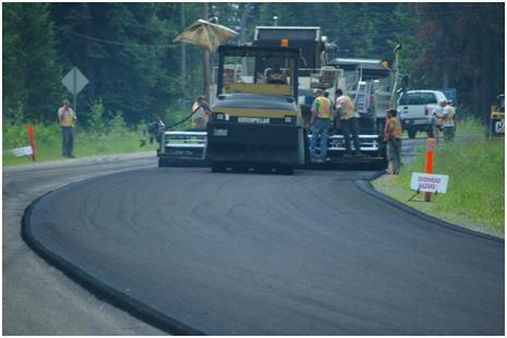 A photo of a construction roller flatting out a freshly asphalted road with several construction workers around it